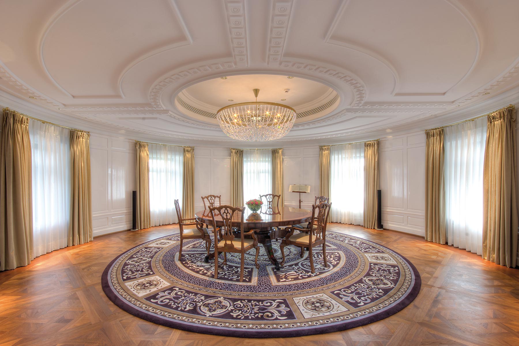 Runder Tisch aus Holz mit Bestuhlung in einem runden Raum, der mit gro&szlig;en Fenstern, sowie wei&szlig;en Vorh&auml;ngen versehen ist als auch mit einem Kronleuchter gestaltet ist