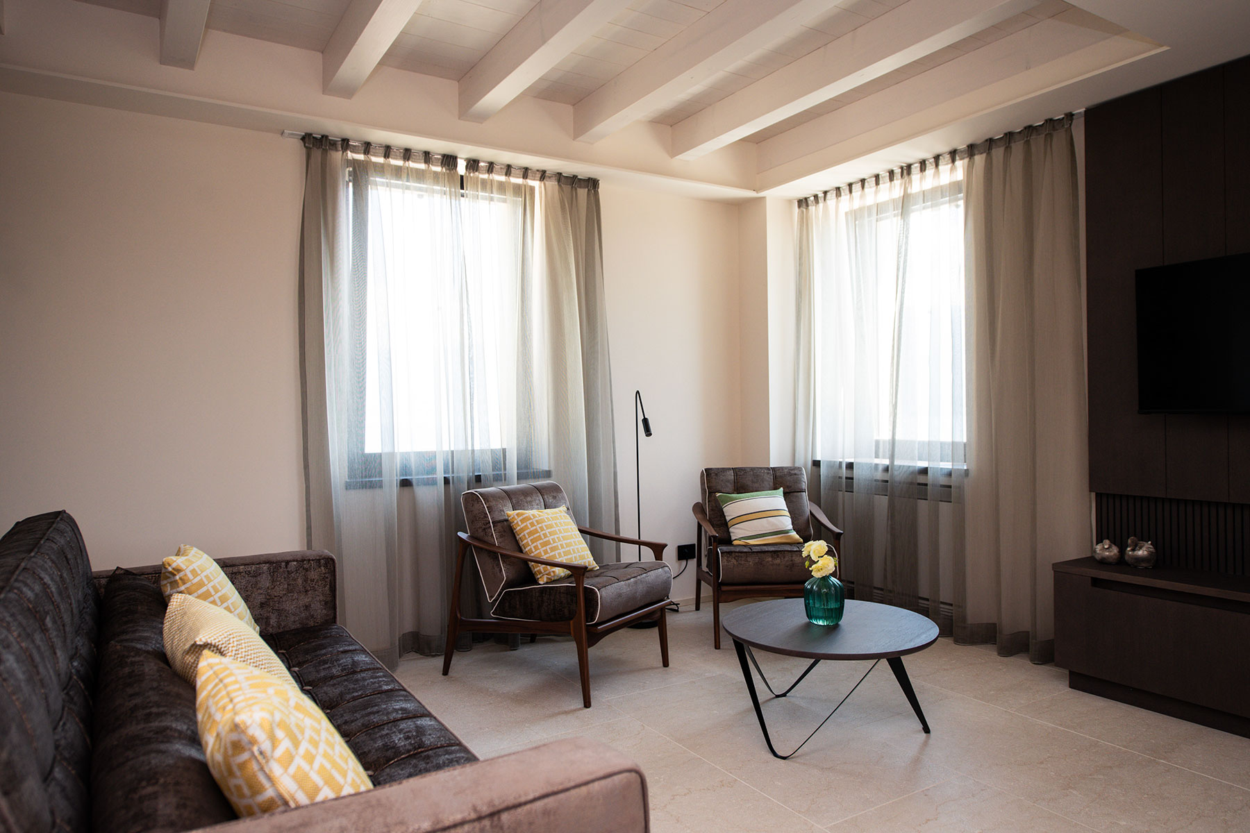 Dunkelbraunes Sofa mit gemusterten gelben Zierkissen und zwei passende Sessel um runden schwarzen Couchtisch vor hellen Fensterfronten im Maison Selva Grand Deluxe Apartment