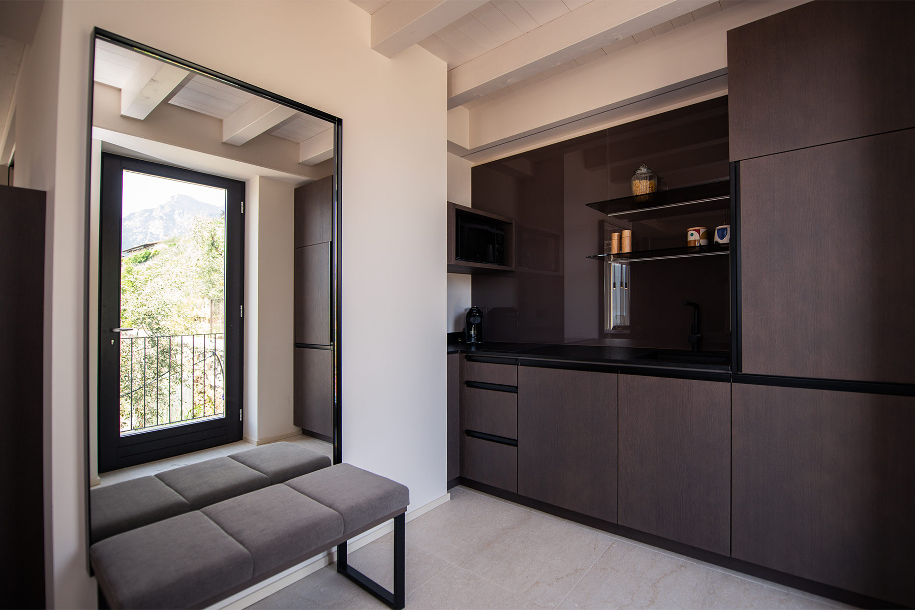 Exklusives Küchendesign im Appia Projekt Gargnano mit grifflosen dunklen Schränken und einer eleganten Spiegelwand die das Tageslicht der gegenüberliegenden Balkontür einfängt