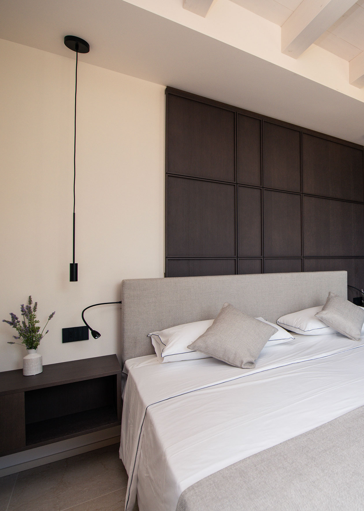 Zentral platziertes Doppelbett mit weißer Bettwäsche und grauen Zierkissen vor raumhoher Wandvertäfelung aus quadratischen dunklen Holzpaneelen im Maison Selva Grand Deluxe Apartment