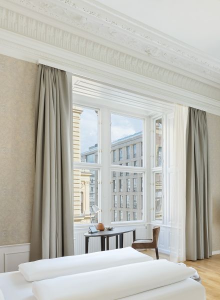 Hell gestaltetes Hotelzimmer im Hotel Rathauspark Wien von Appia mit Fokus auf die hohen Fensterfronten und die prachtvollen weißen Stuckelemente an der Decke