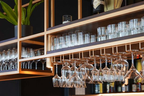Detail der Hotelbar mit wandhängendem Regal für Flaschen und Gläser Metallrahmen warm hinterlegter Holzstruktur und integrierter Beleuchtung