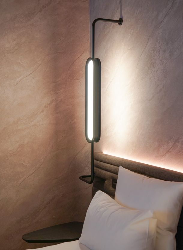 Detail aus dem Hotelzimmer mit weichem Kopfteil warm hinterleuchteter Wandstruktur und schlanker Leuchte seitlich am Bett