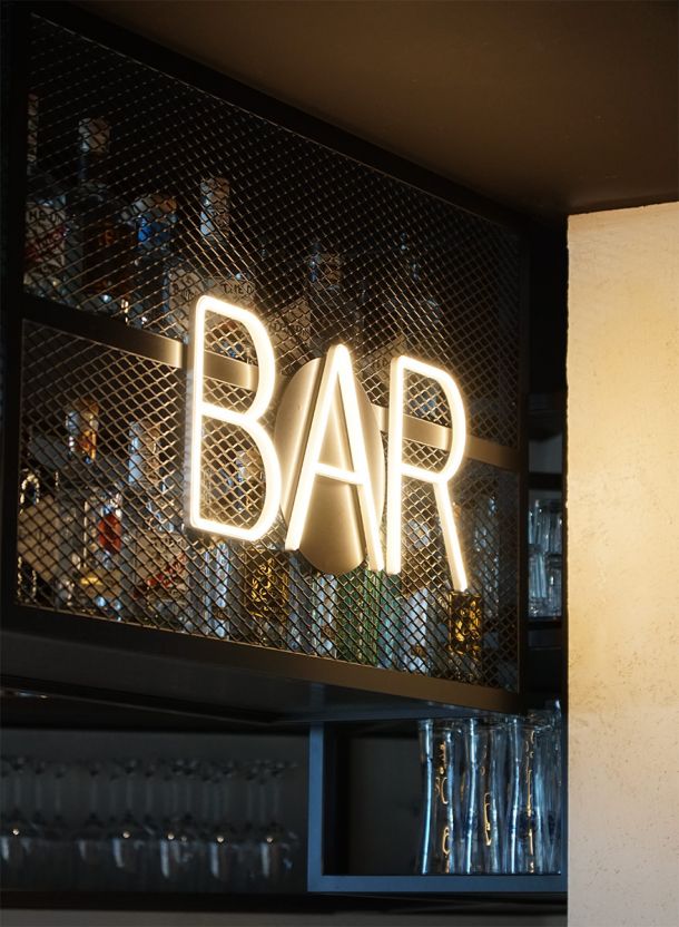 Bar Leuchtschild im DEIN APART Unterföhring mit großem beleuchtetem Schriftzug BAR vor schwarzem Metallgitter und Flaschenregal