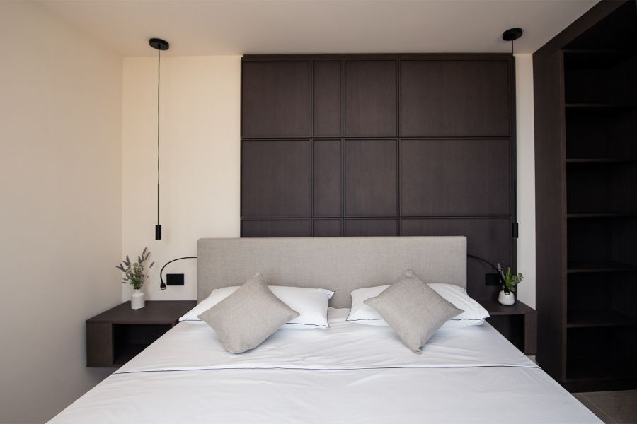 Zentral platziertes Doppelbett mit weißer Bettwäsche und grauen Zierkissen vor raumhoher Wandvertäfelung aus quadratischen dunklen Holzpaneelen im Maison Selva Grand Deluxe Apartment