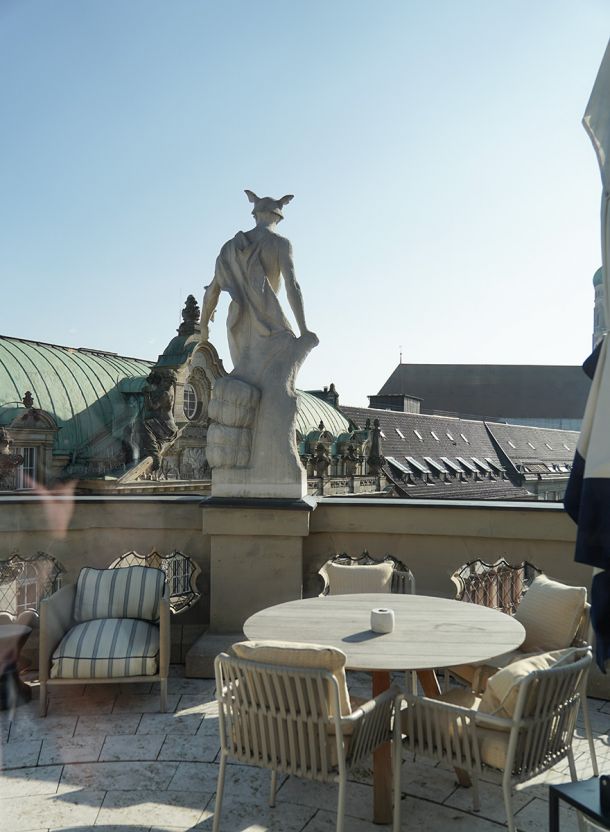 Aussichtsterrasse im Rosewood München mit steinernem Sonnendeck und einer Sitzgruppe aus hellem Geflecht rundem Tisch und gepolsterten Sesseln