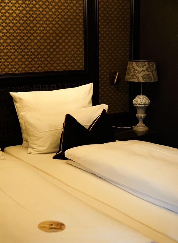 Atmosphärischer Bettbereich im Gasthof Schäfflerwirt mit hellem Bett dunklem Wandfeld ornamentalen Stuckrahmen und dekorativer Tischleuchte auf dem Nachttisch