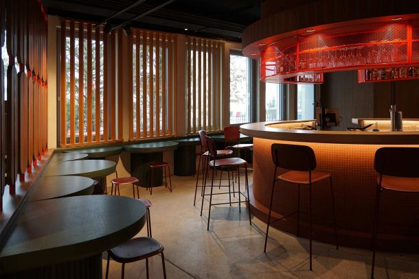 Zentraler Barbereich mit mosaikverkleidetem Tresen warmer Rotlichtbeleuchtung vertikalen Holzlamellen und Sitzplätzen entlang der Fensterfront