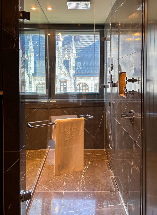 Badezimmer im Do & Co Hotel München mit bodengleicher Dusche großformatigem Marmorboden Glasabtrennung und direktem Blick auf den Marienplatz