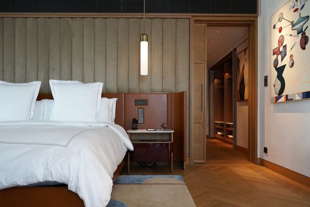 Blick durchs Doppelzimmer mit hohem Kopfteil vertikaler Wandstruktur schmaler Pendelleuchte warmem Holzboden und offener Verbindung zum Vorraum